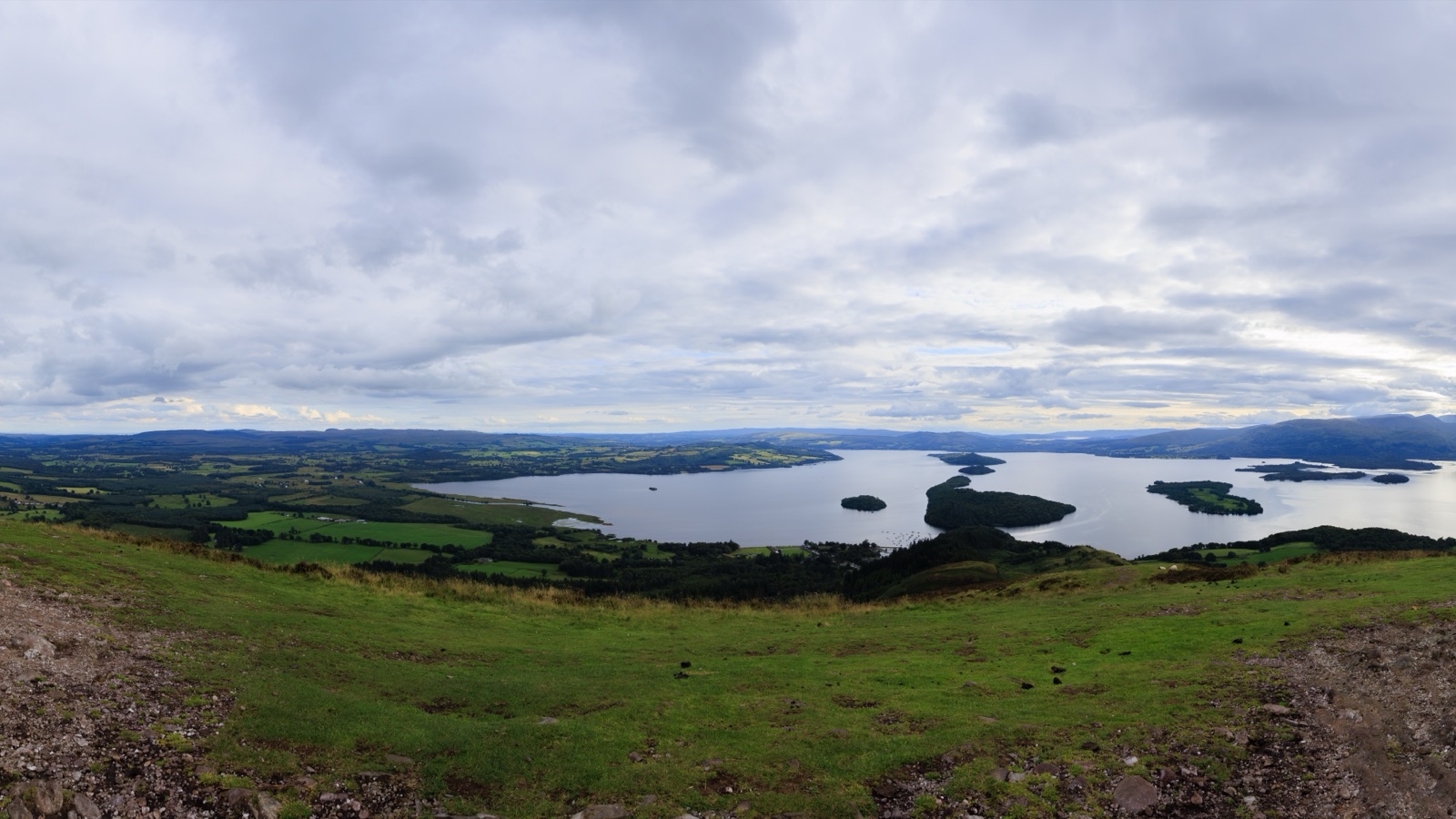 Elevated panorama from Conic Hill across Loch Lomond and its wooded islands under a cloudy sky.